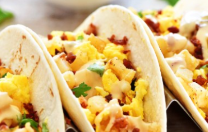 Two warm flour tortillas filled with fluffy scrambled eggs, crispy bacon, and dripping with creamy hot queso on a wooden board.