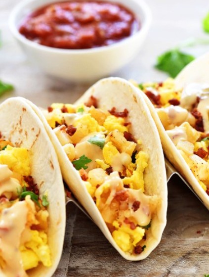 Two warm flour tortillas filled with fluffy scrambled eggs, crispy bacon, and dripping with creamy hot queso on a wooden board.