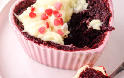 A vibrant red velvet mug cake topped with creamy frosting and heart-shaped sprinkles, served in a white ceramic mug