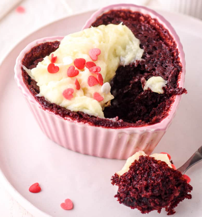 A vibrant red velvet mug cake topped with creamy frosting and heart-shaped sprinkles, served in a white ceramic mug