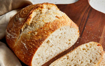 A golden-brown, crusty loaf of artisan sourdough bread with a beautiful decorative score on top, sitting on a floured surface.
