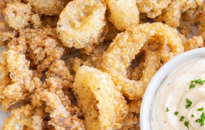 A plate of golden, crispy fried calamari rings served with fresh lemon wedges and a small bowl of creamy garlic aioli dip.