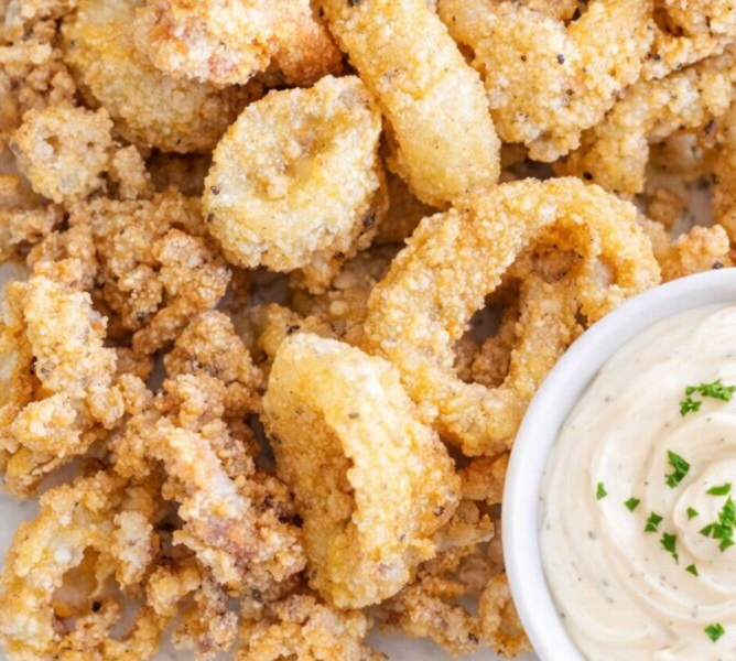 A plate of golden, crispy fried calamari rings served with fresh lemon wedges and a small bowl of creamy garlic aioli dip.