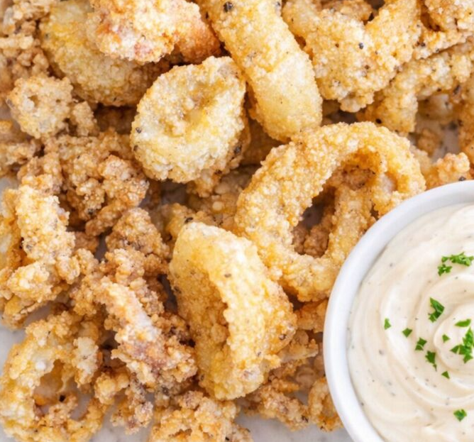 A plate of golden, crispy fried calamari rings served with fresh lemon wedges and a small bowl of creamy garlic aioli dip.