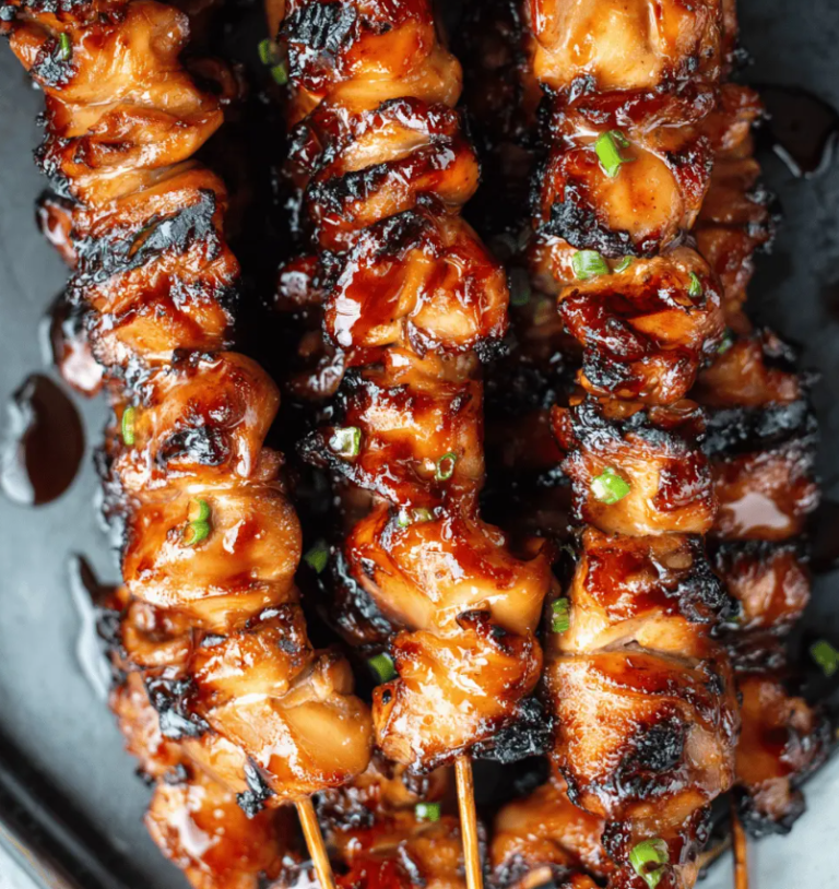 A platter of glossy, caramelized Filipino barbecue chicken skewers garnished with fresh scallions and charred lemon halves.