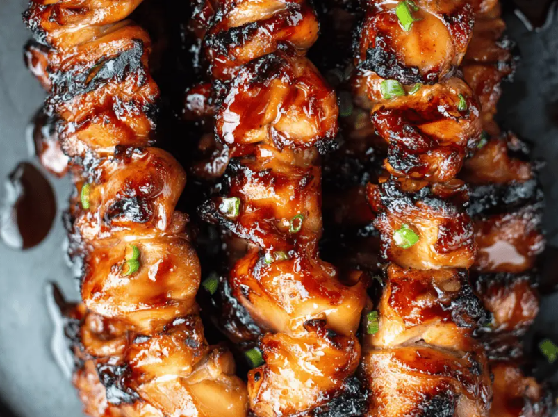 A platter of glossy, caramelized Filipino barbecue chicken skewers garnished with fresh scallions and charred lemon halves.