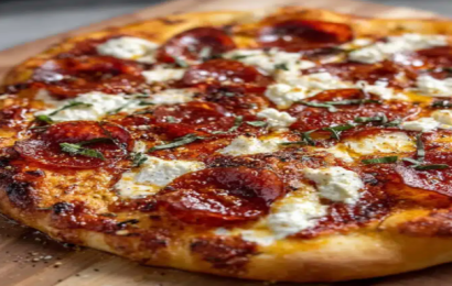 A freshly baked pepperoni pizza topped with dollops of creamy ricotta cheese and a shiny drizzle of hot honey on a wooden pizza peel.