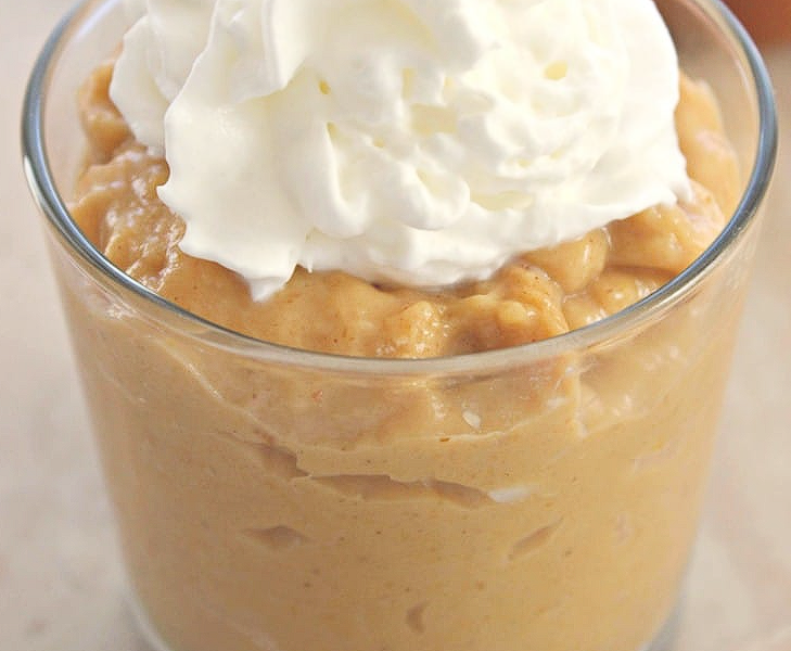A close-up of a rich, creamy bowl of homemade spiced pumpkin pudding, generously topped with a dollop of fresh whipped cream and a sprinkle of cinnamon on a rustic wooden table.