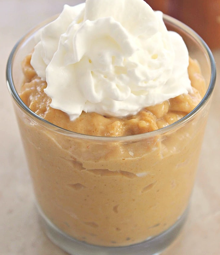 A close-up of a rich, creamy bowl of homemade spiced pumpkin pudding, generously topped with a dollop of fresh whipped cream and a sprinkle of cinnamon on a rustic wooden table.