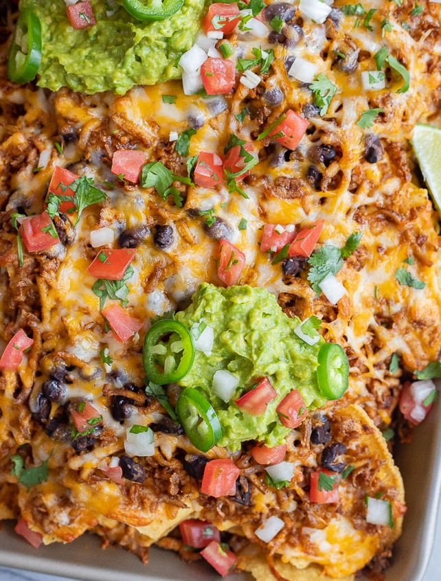 A large sheet pan piled high with crispy tortilla chips, melted cheese, seasoned shredded tofu, fresh jalapeños, and guacamole.