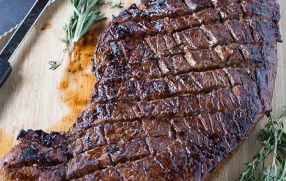 Thinly sliced grilled London broil steak with charred grill marks, served with a side of fresh herb butter on a wooden carving board.