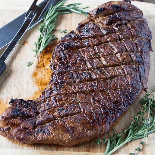 Thinly sliced grilled London broil steak with charred grill marks, served with a side of fresh herb butter on a wooden carving board.