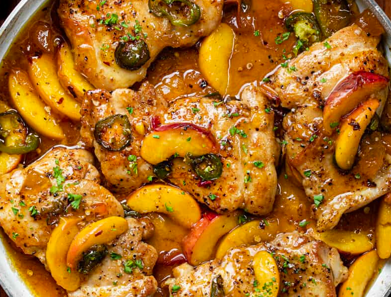 A hot cast-iron skillet featuring juicy pan-seared chicken breasts smothered in a glossy, golden peach glaze with sliced green jalapeños and fresh cilantro.