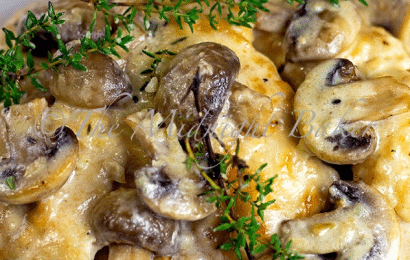 A skillet filled with tender chicken breasts smothered in a rich, creamy brown mushroom sauce and melted Asiago cheese, garnished with fresh thyme.