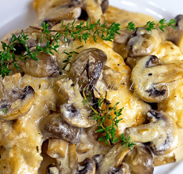 A skillet filled with tender chicken breasts smothered in a rich, creamy brown mushroom sauce and melted Asiago cheese, garnished with fresh thyme.