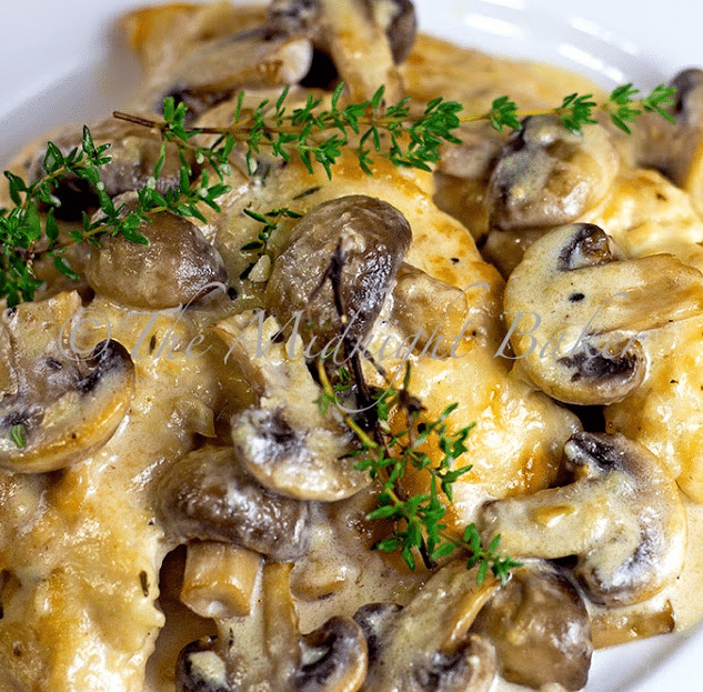 A skillet filled with tender chicken breasts smothered in a rich, creamy brown mushroom sauce and melted Asiago cheese, garnished with fresh thyme.