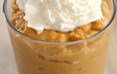 Creamy, spiced homemade pumpkin pudding served in elegant glass jars, topped with a generous dollop of fresh whipped cream and a sprinkle of cinnamon on a rustic wooden board.