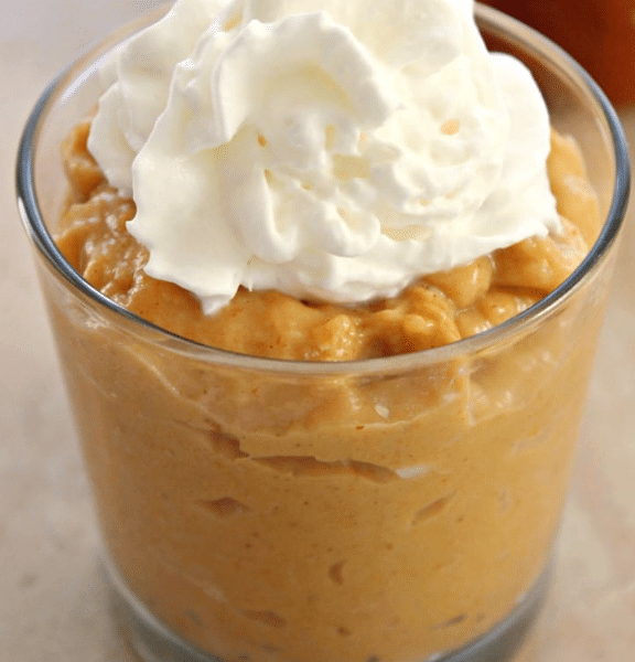 Creamy, spiced homemade pumpkin pudding served in elegant glass jars, topped with a generous dollop of fresh whipped cream and a sprinkle of cinnamon on a rustic wooden board.