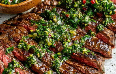 A perfectly grilled flank steak sliced against the grain on a wooden board, generously topped with a vibrant, bright green garlic and herb homemade chimichurri sauce.